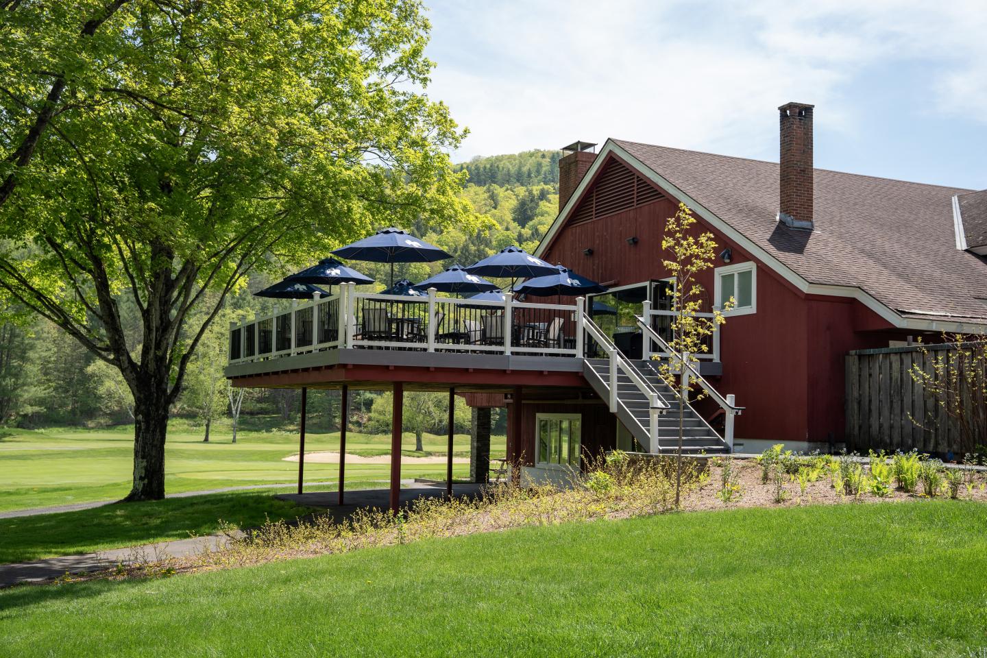Red house with deck and blue umbrellas, set in a green, sunny landscape.