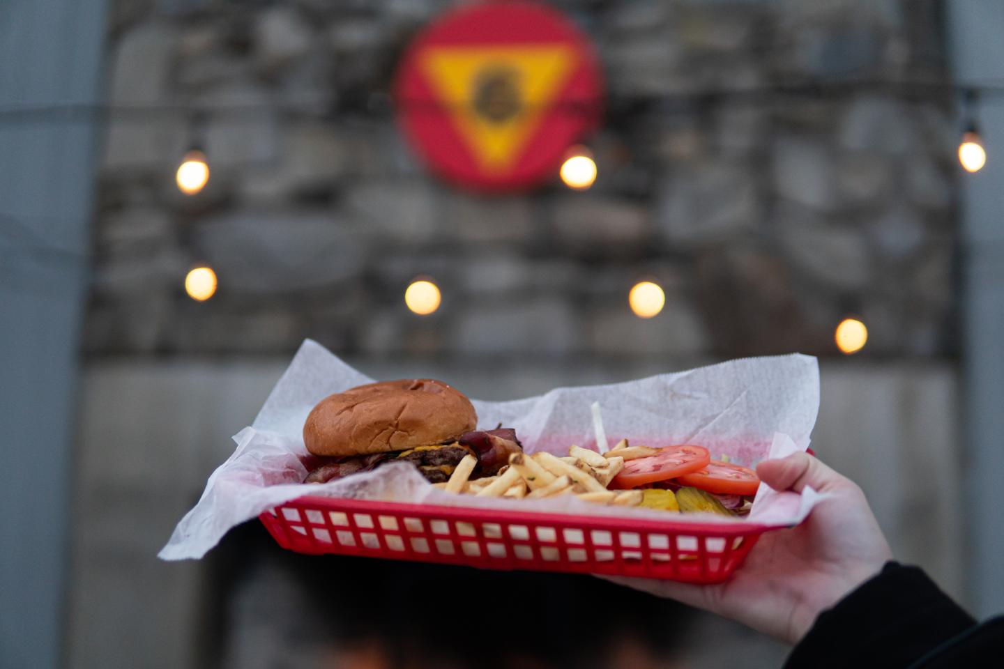 Burger and fries in a red basket held up against a gray brick wall with lights.