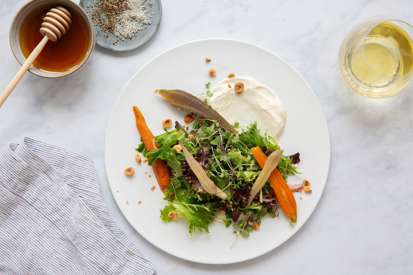 Salad with greens, carrots, and dressing on a white plate, next to honey and wine.