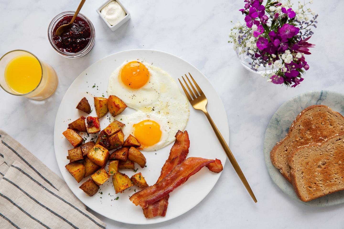 Breakfast with eggs, potatoes, bacon, toast, and orange juice on a marble table.