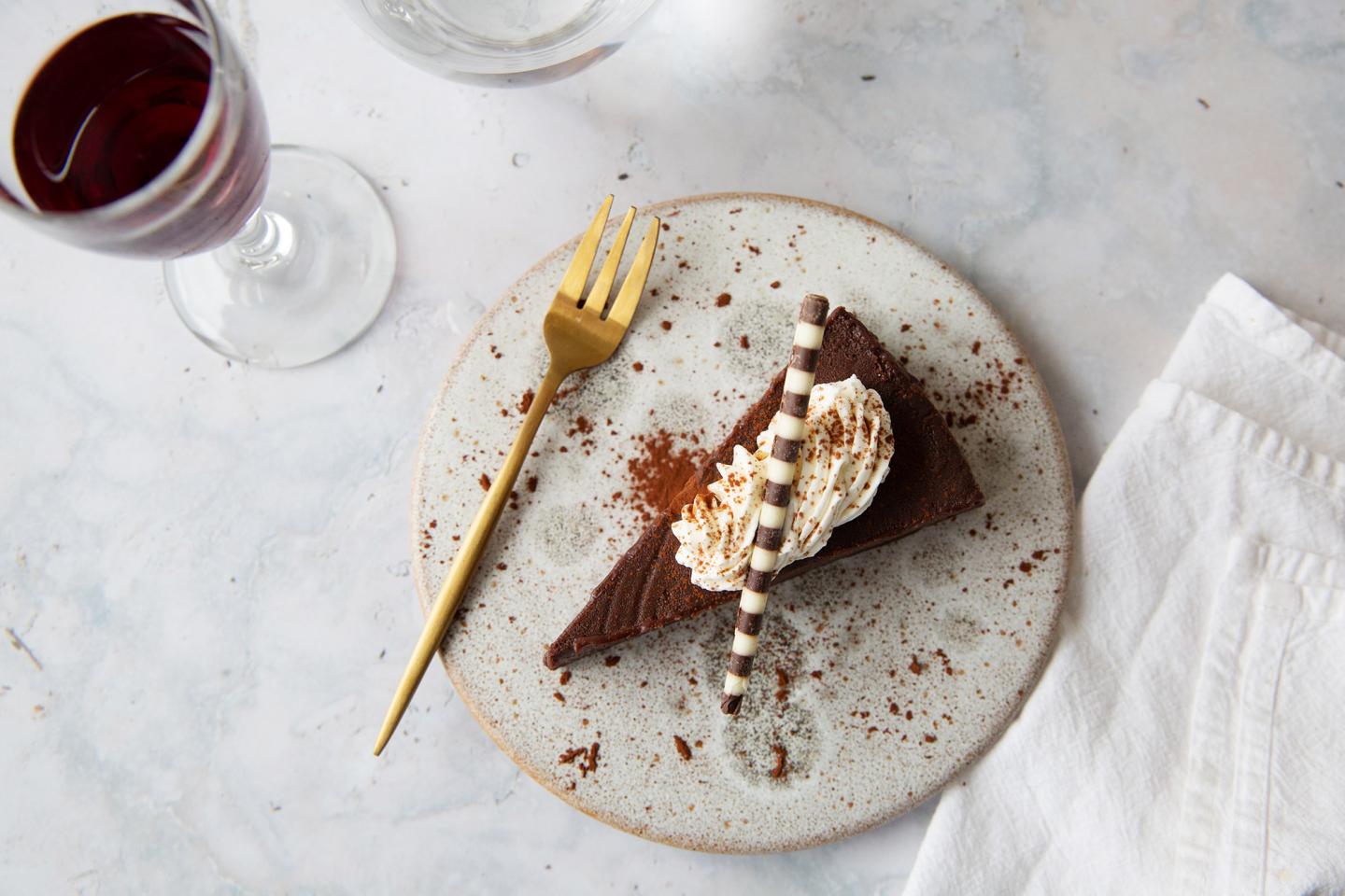 Chocolate cake slice with cream on a plate, next to a glass of red wine and a gold fork.