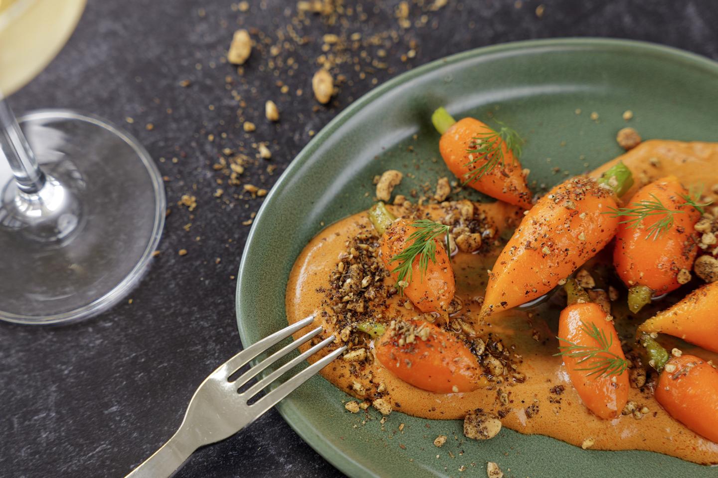 Carrots on a green plate with sauce, crumbs, and a fork beside a glass of white wine.