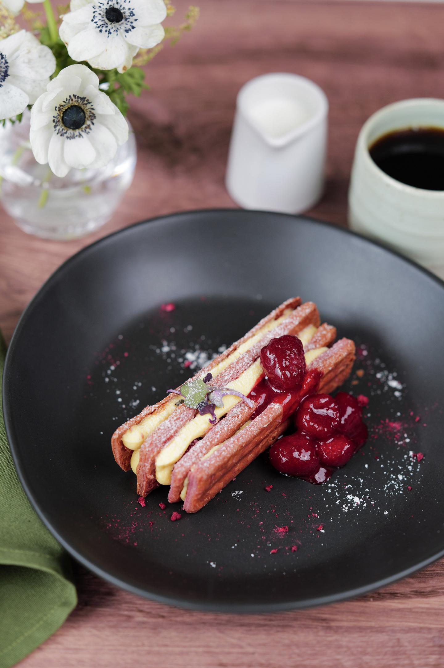 Cherries and cream sandwich on a black plate, with coffee and flowers nearby.