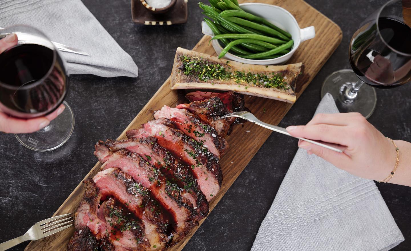 Sliced steak on a wooden board with wine glasses and green beans.