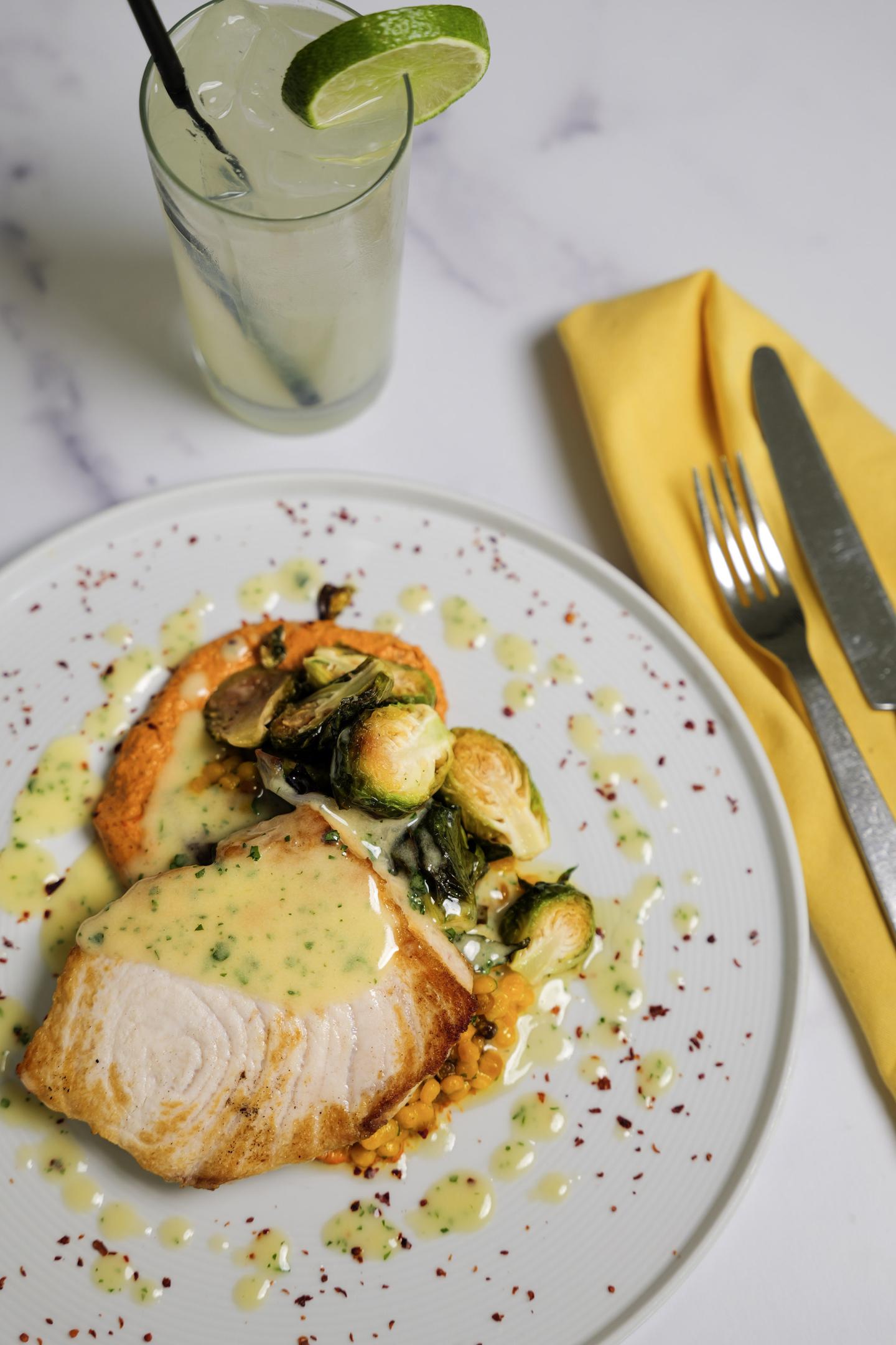Grilled fish with Brussels sprouts and sauce, beside lemonade and utensils on a table.