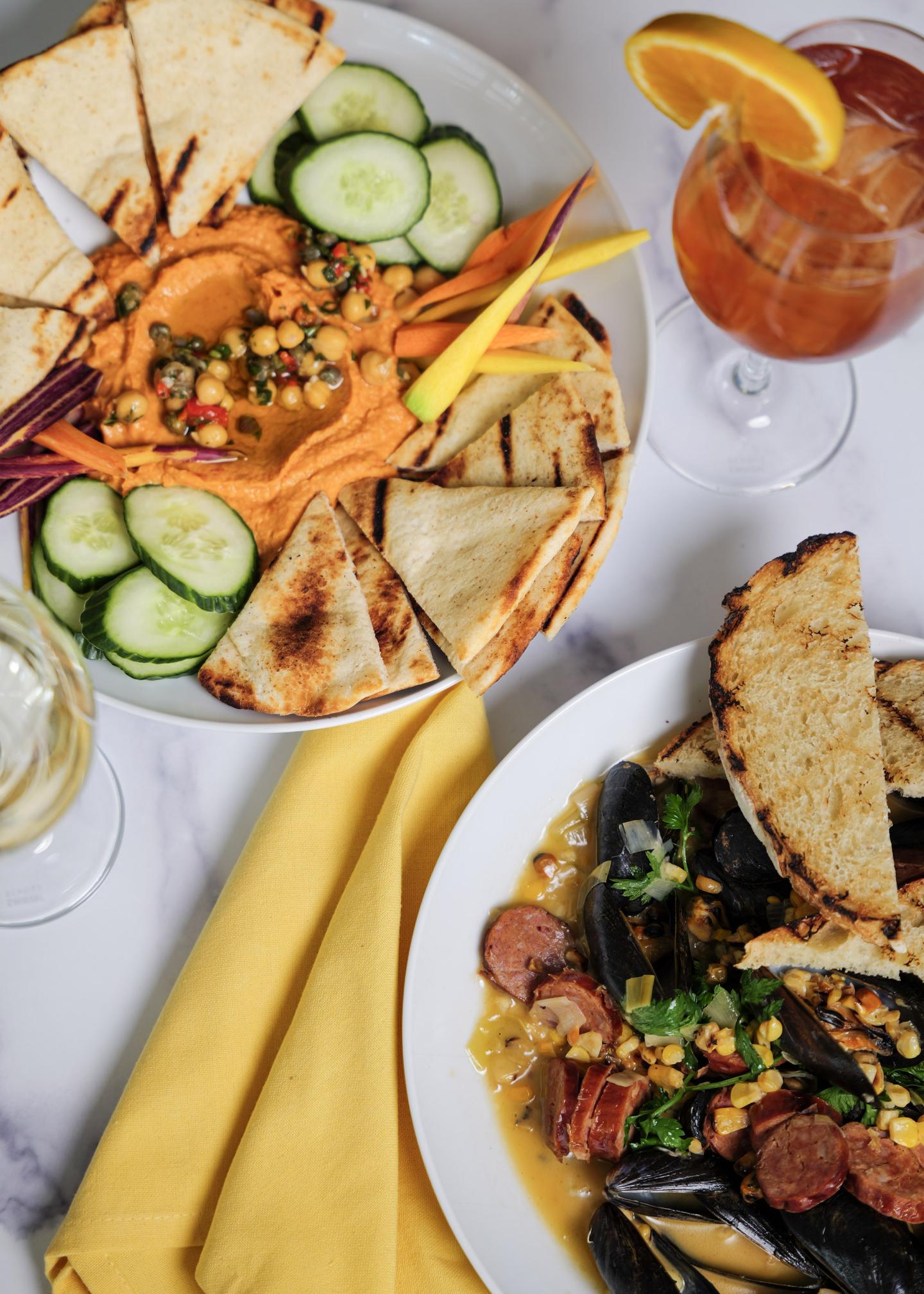 Colorful hummus platter with vegetables and seafood dish, accompanied by drinks.