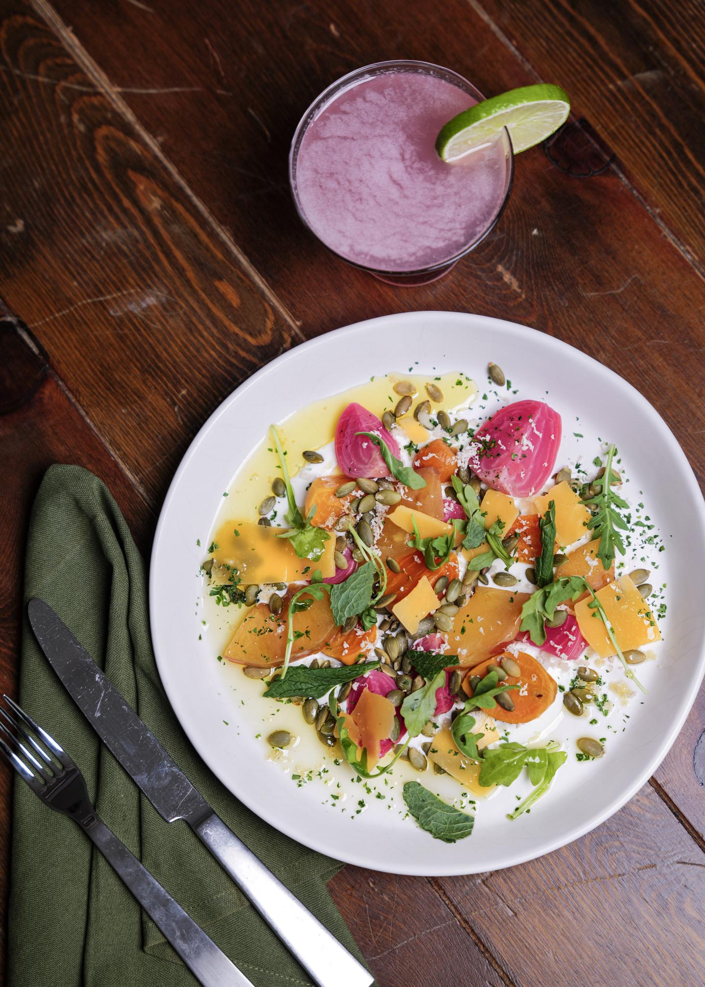 Salad with vibrant vegetables on a white plate, next to a purple drink with a lime slice.