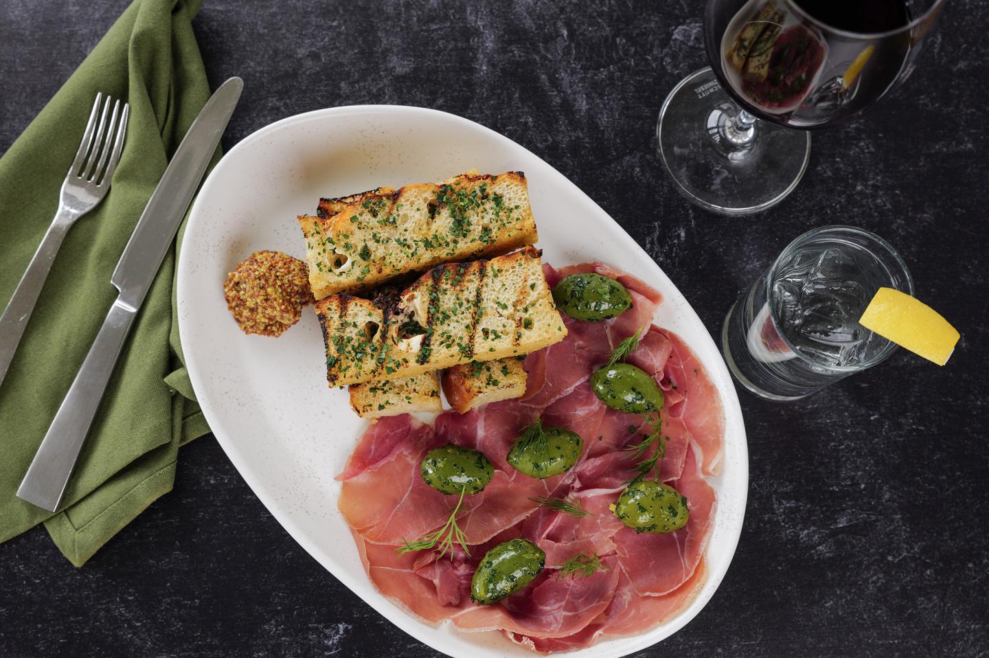 Sliced bread, prosciutto, and greens on a white plate, with wine and lemon water nearby.