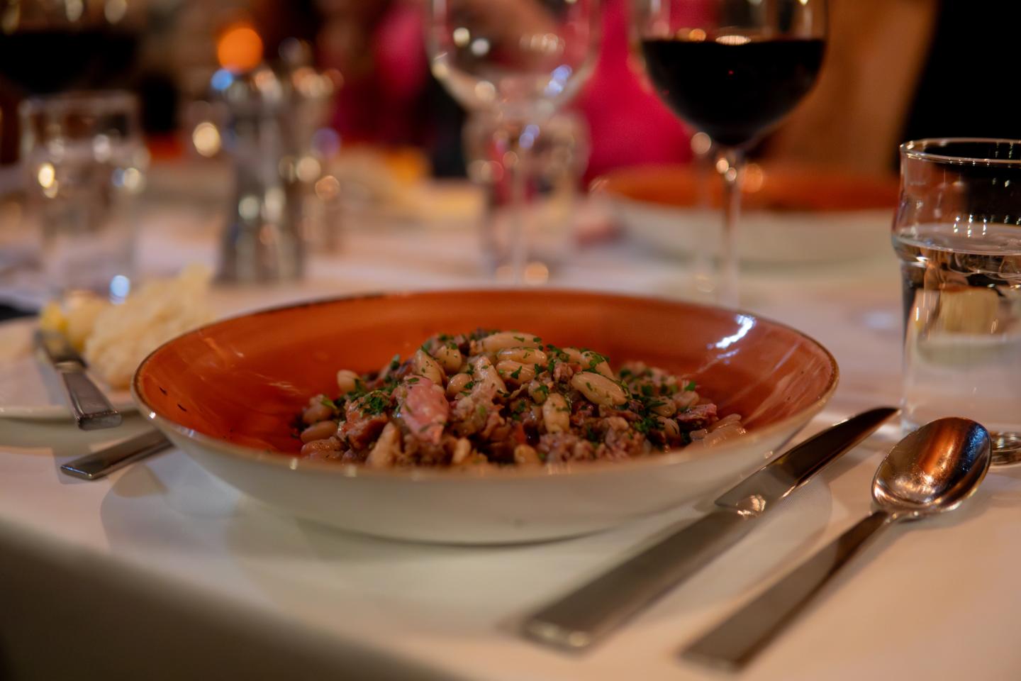 Pasta dish in a red bowl on a dining table with glasses and utensils.
