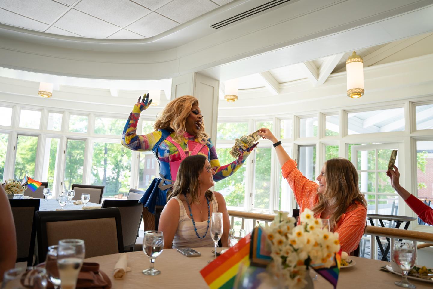Drag performer entertaining a seated group at a bright, festive event with Pride flags.