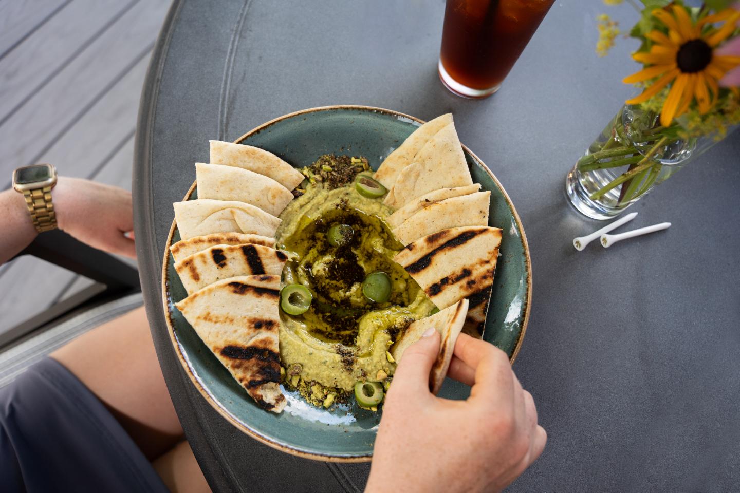 Pita bread with dip and olives on a green plate, next to iced drink and flowers.