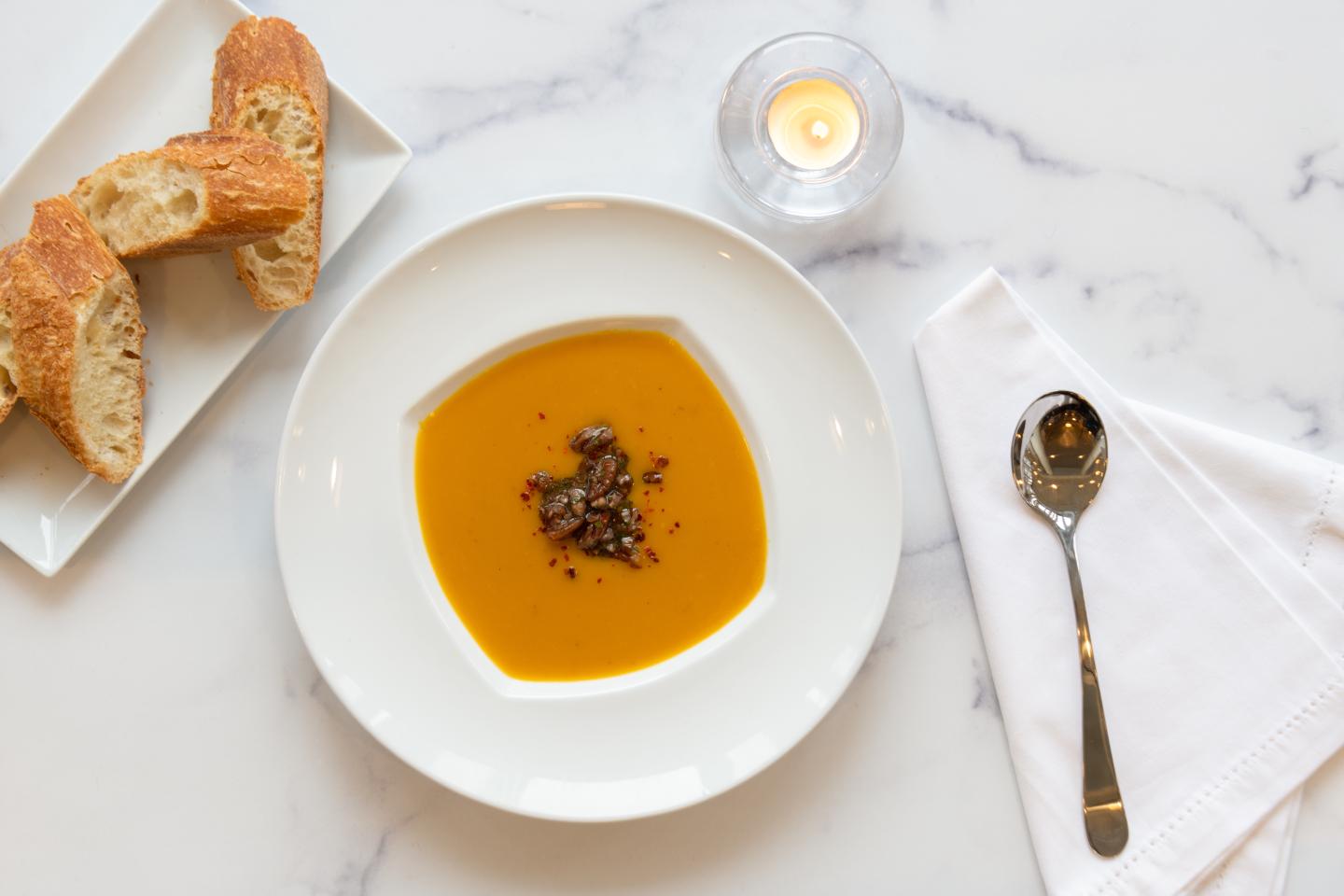 Bowl of orange soup with herbs, bread on side, spoon and napkin nearby.