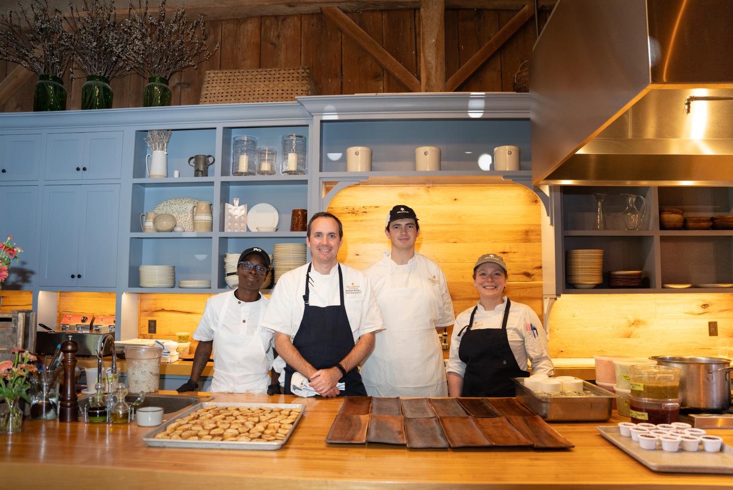 Four chefs in a rustic kitchen with wooden shelves and blue cabinets.