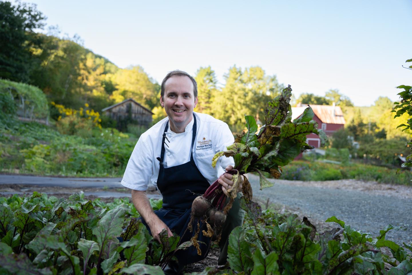 Chef in a garden holding freshly picked vegetables, with a barn in the background.