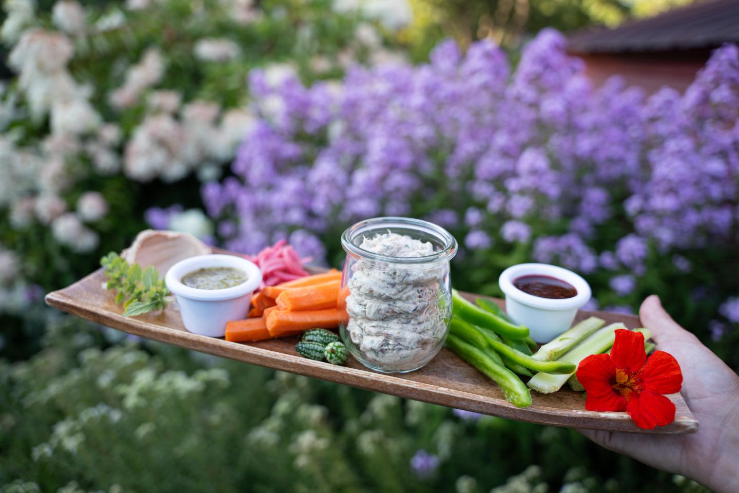 Wooden tray with dips, veggies, and flowers against a garden backdrop.