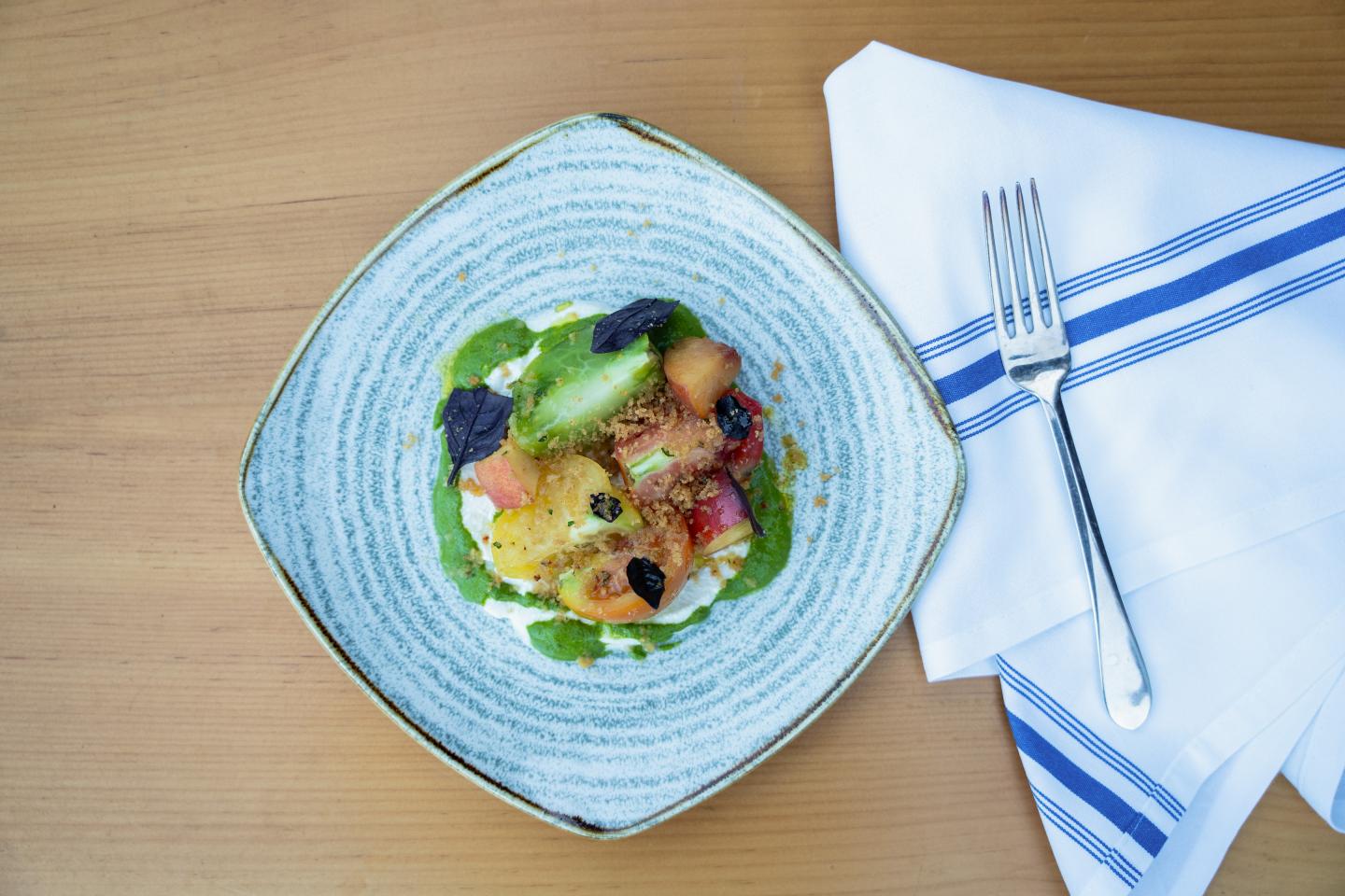Elegant dish with vegetables and sauce on a blue plate, next to a napkin and fork.