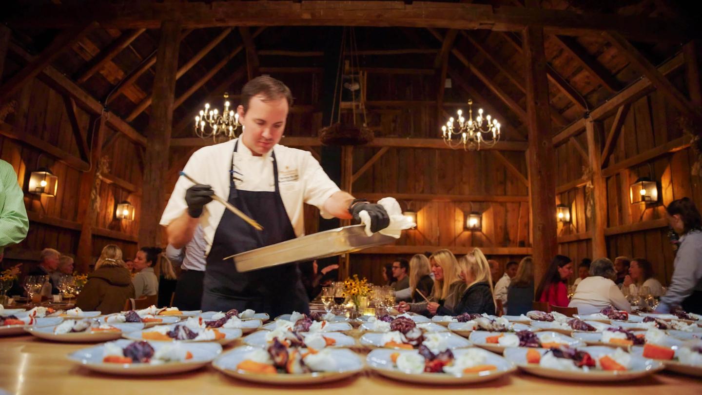 Chef plating dishes in a rustic, warmly lit banquet hall.