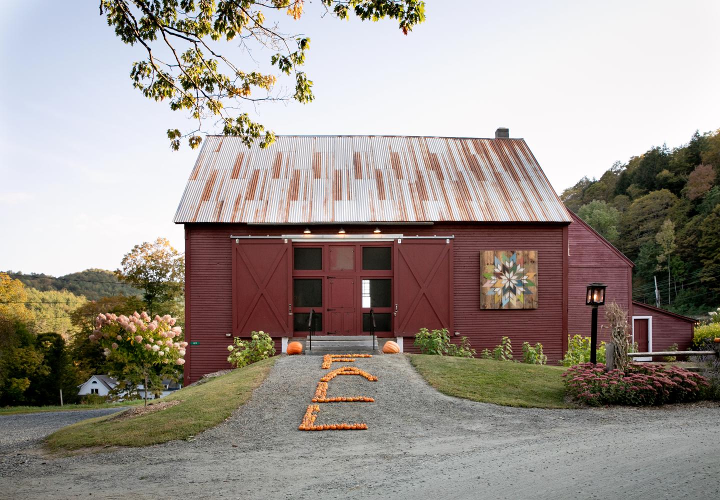 Red barn with a rusted roof on a gravel path, surrounded by autumn trees.