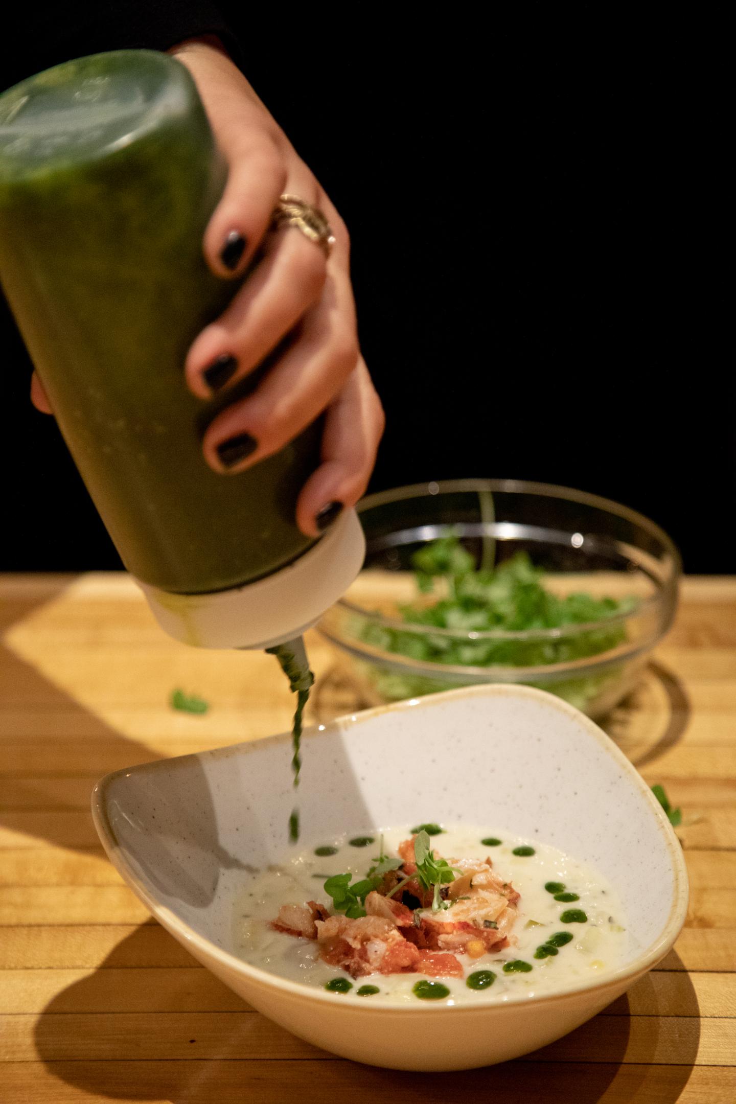 Hand drizzling green sauce over soup in a white bowl with bacon pieces.