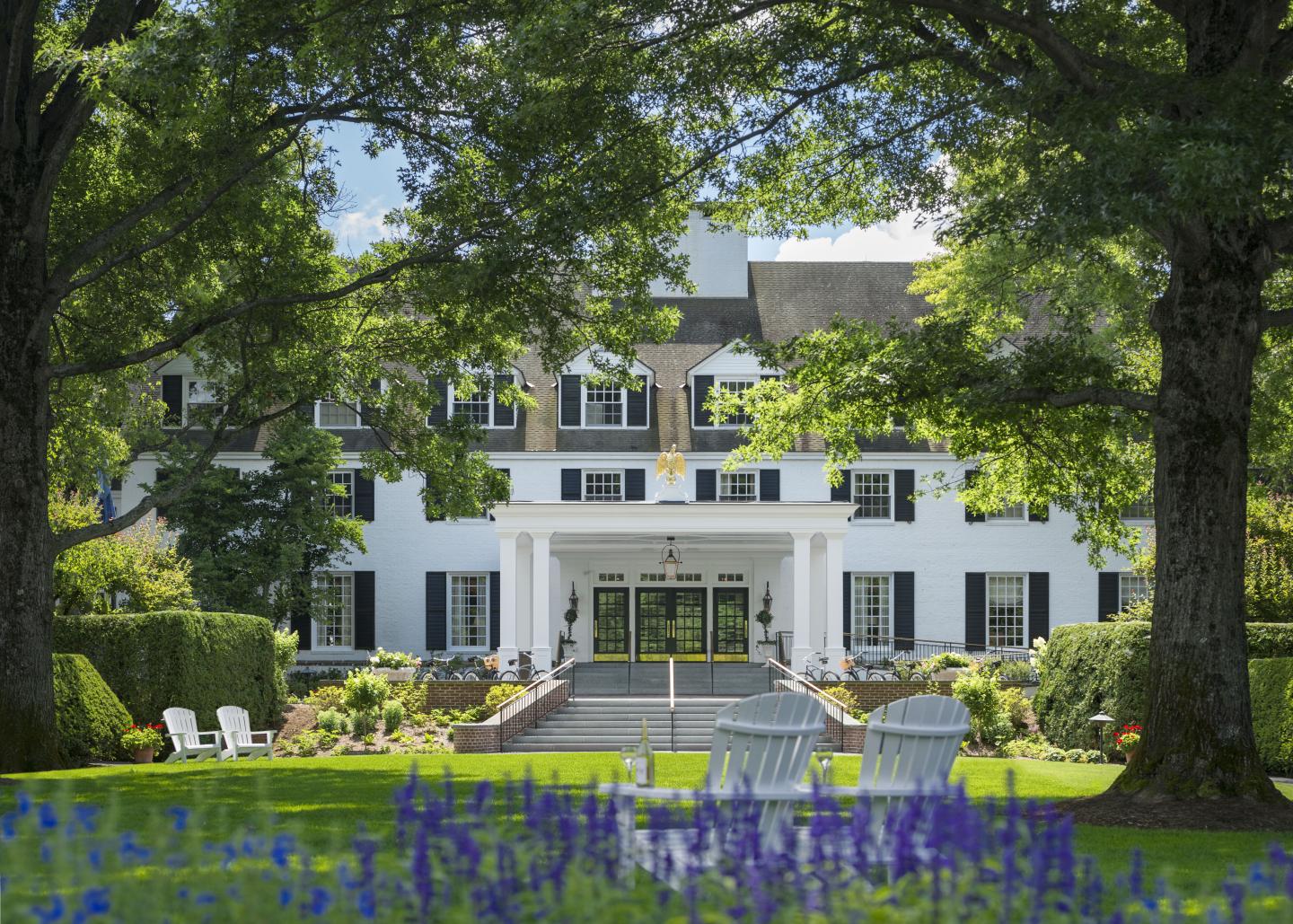 Elegant white mansion with columns, surrounded by lush trees and garden chairs.