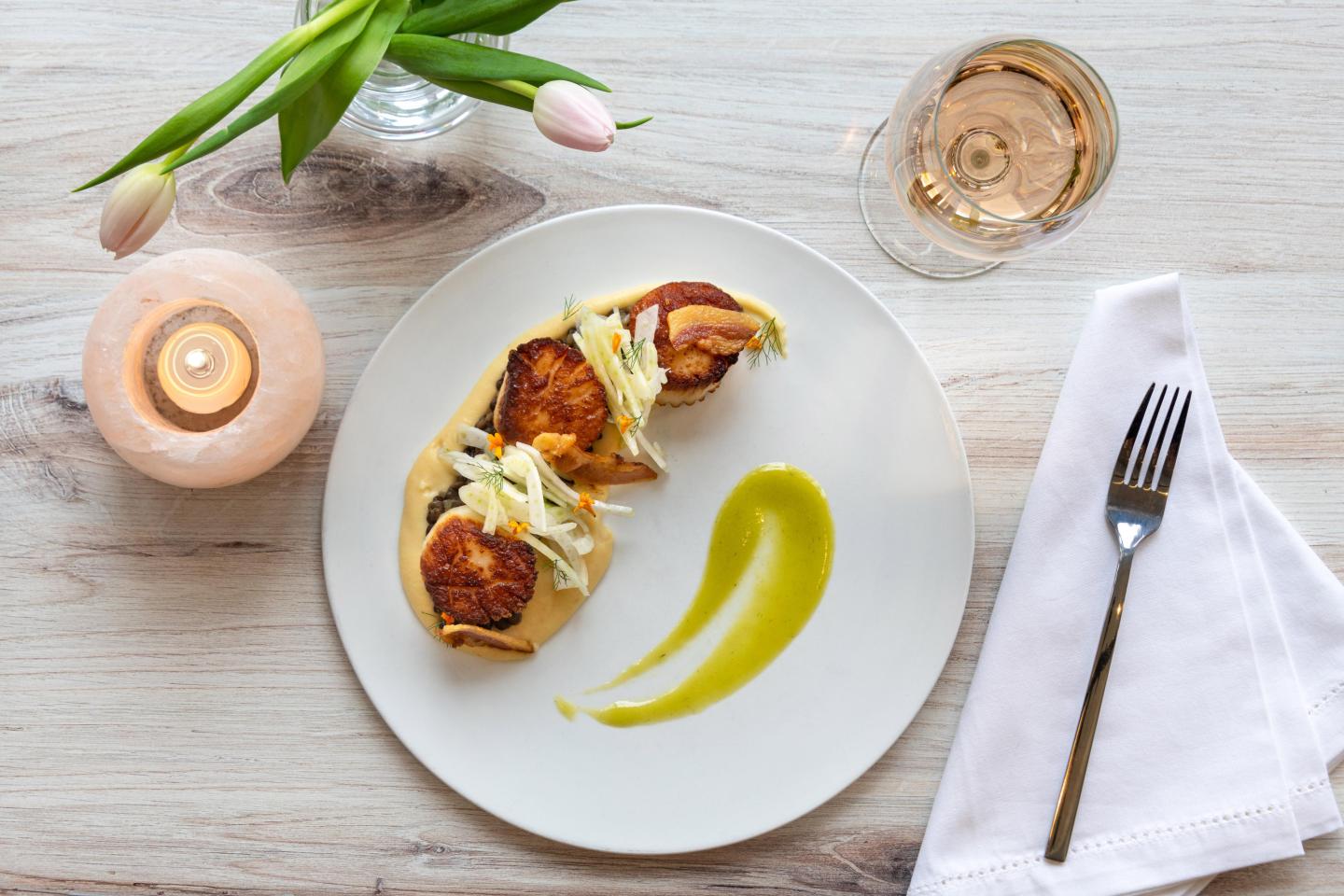 Scallops with garnish on a white plate, next to a candle, tulips, and a glass of wine.