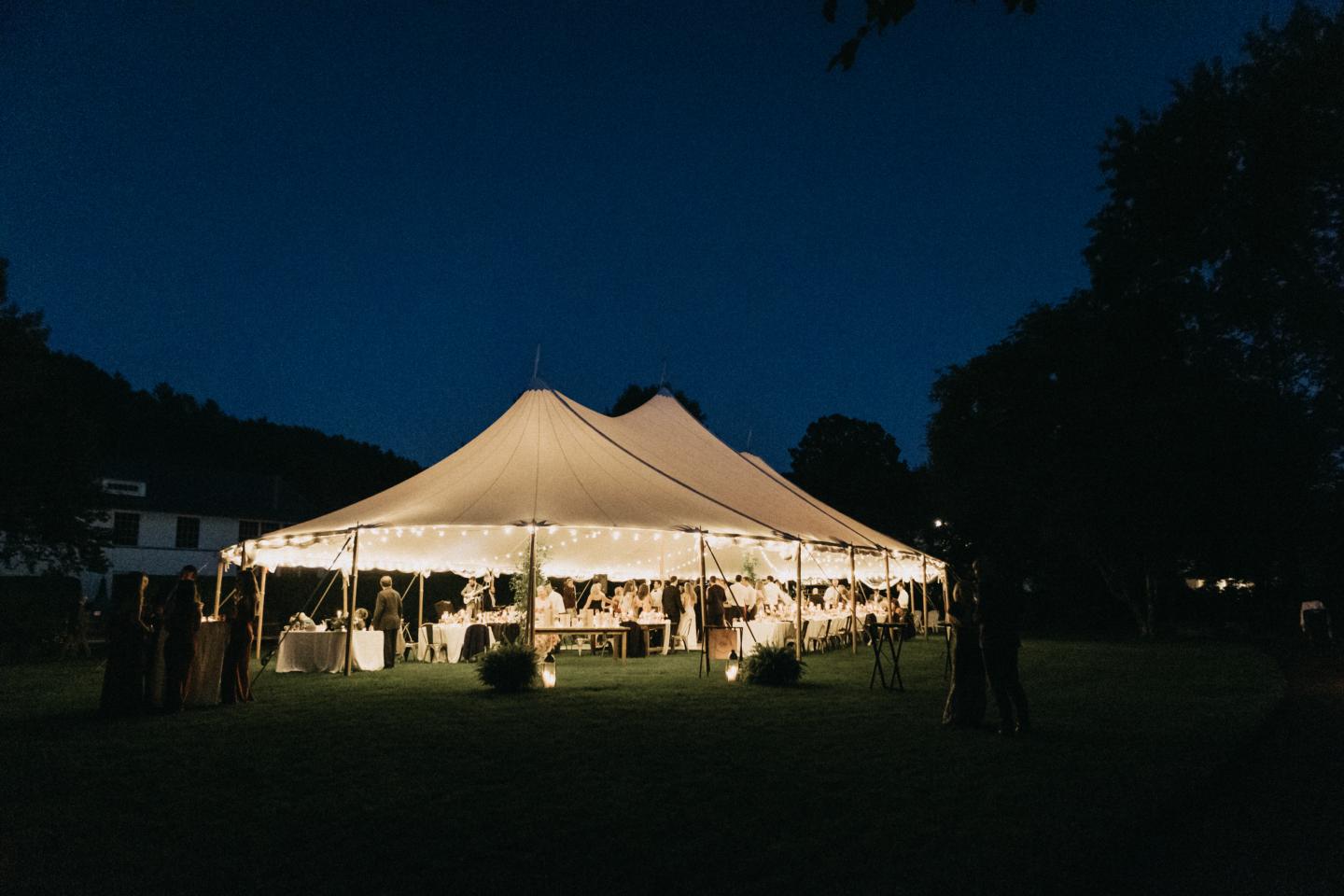 Large white tent illuminated at night, surrounded by trees and guests.