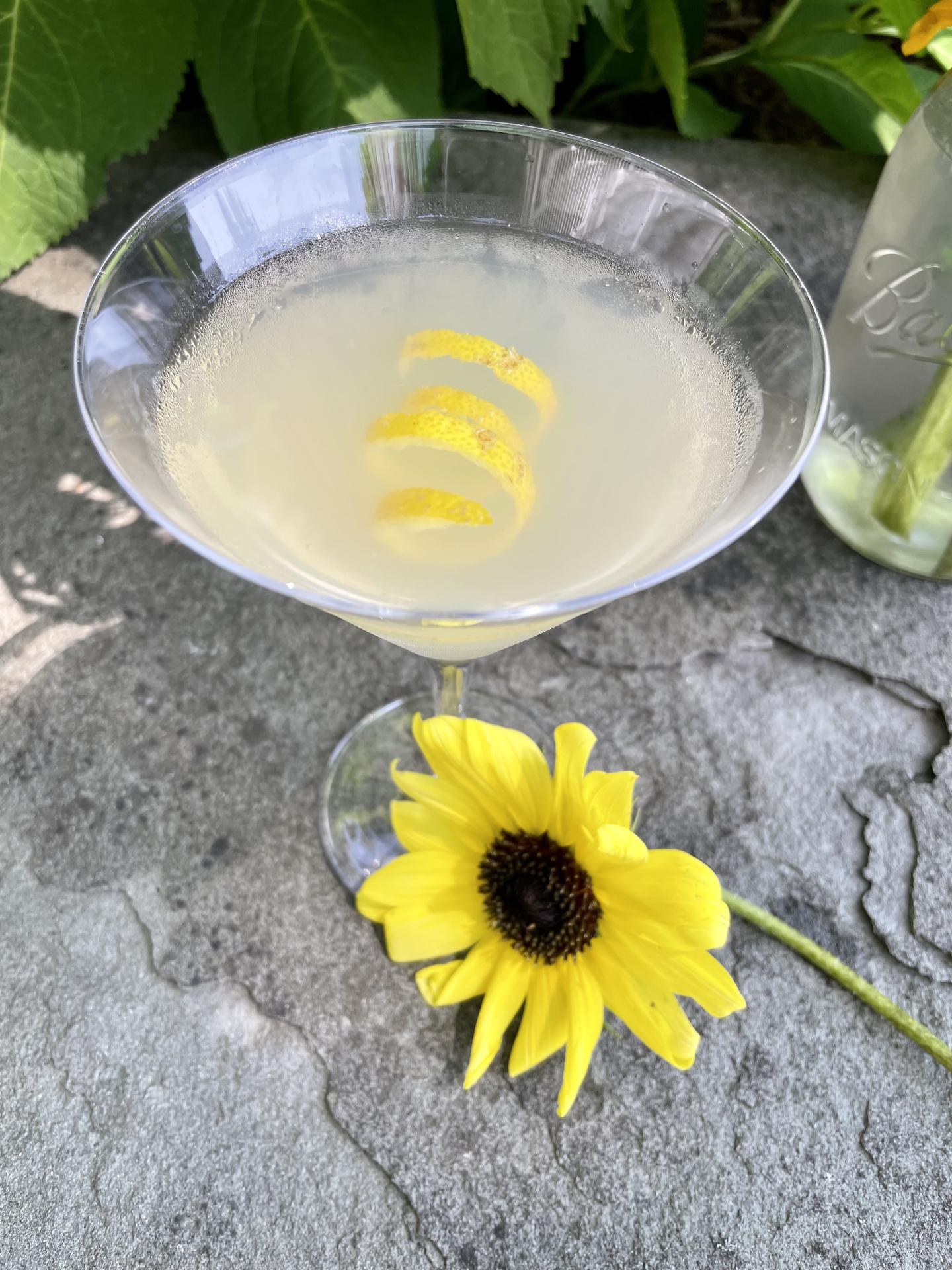 Cocktail in a martini glass with lemon twist, next to a yellow flower on a stone surface.