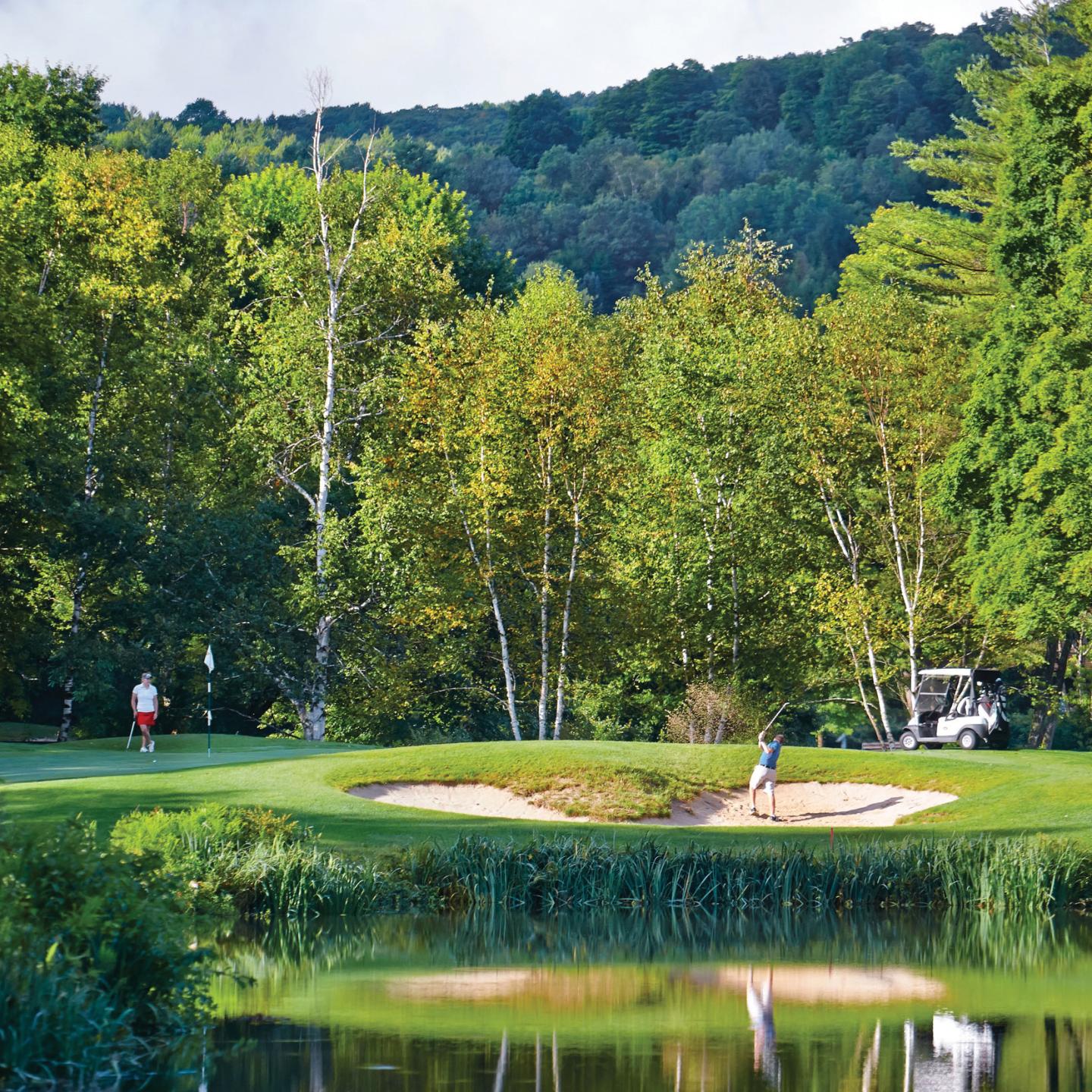 Golfers on a lush green course beside a lake, surrounded by trees and hills.