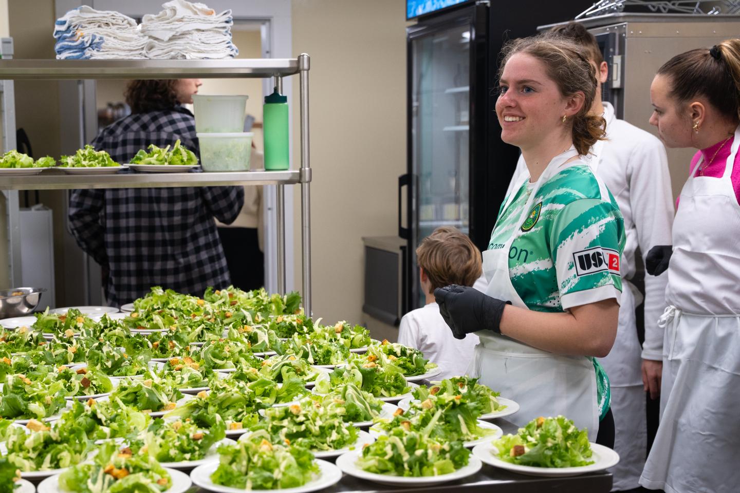 Cooks preparing plates of salad in a kitchen, smiling and wearing aprons.