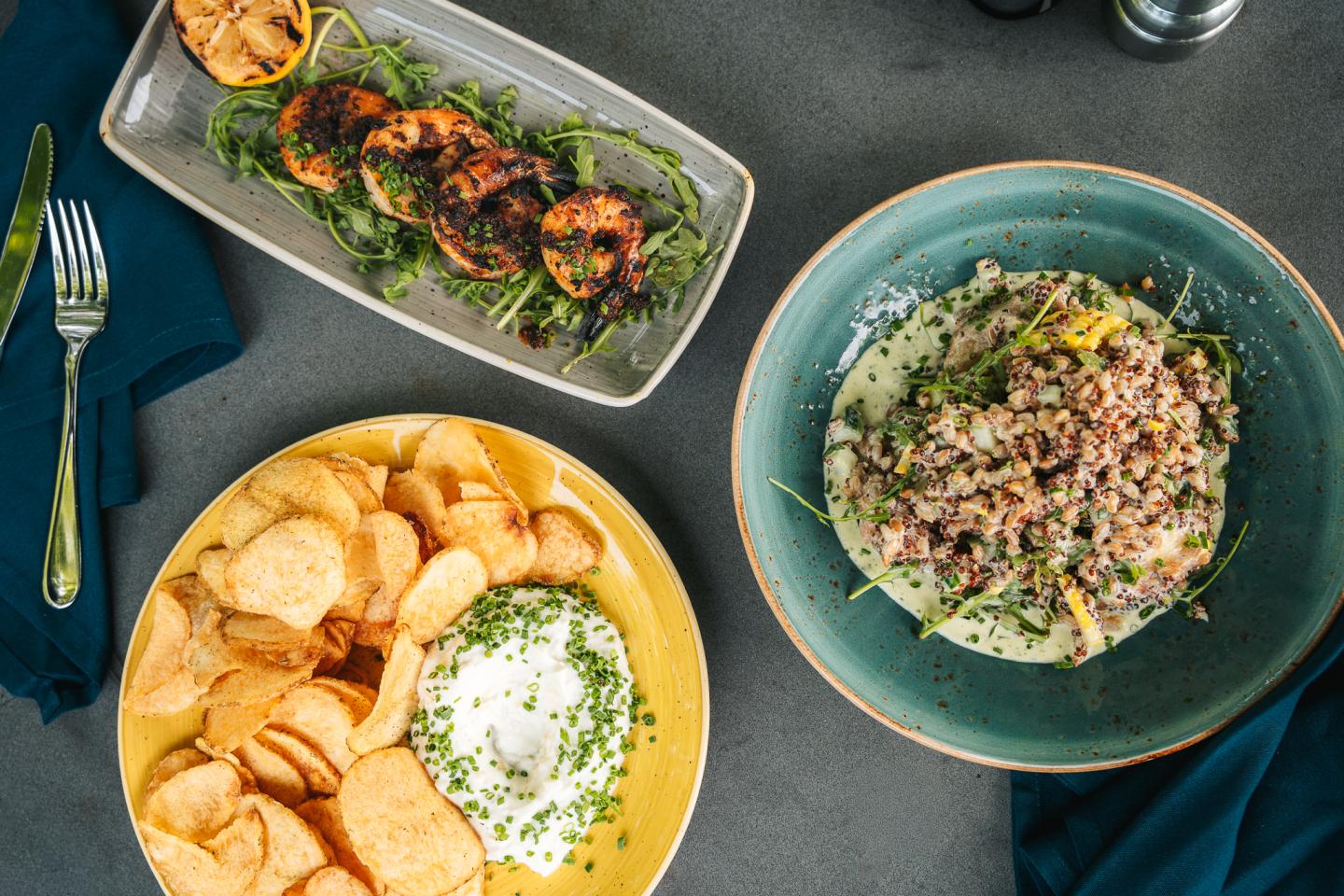 Grilled shrimp skewers, chips with dip, and a grain salad on a table.