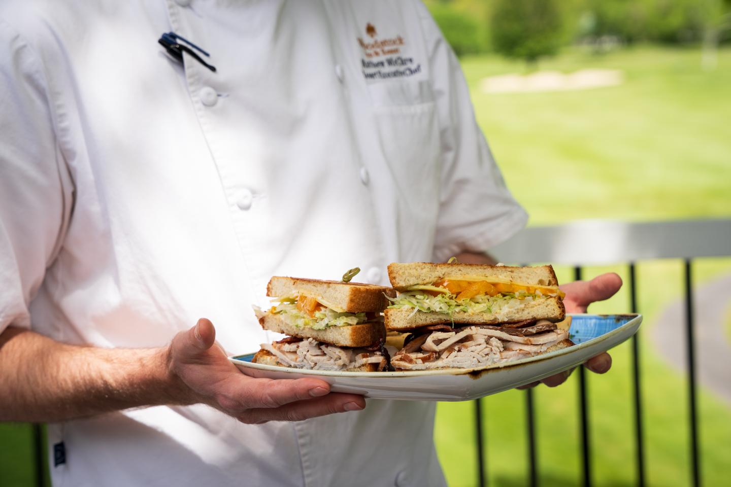 Chef holding a plate with sandwiches on a sunny patio.