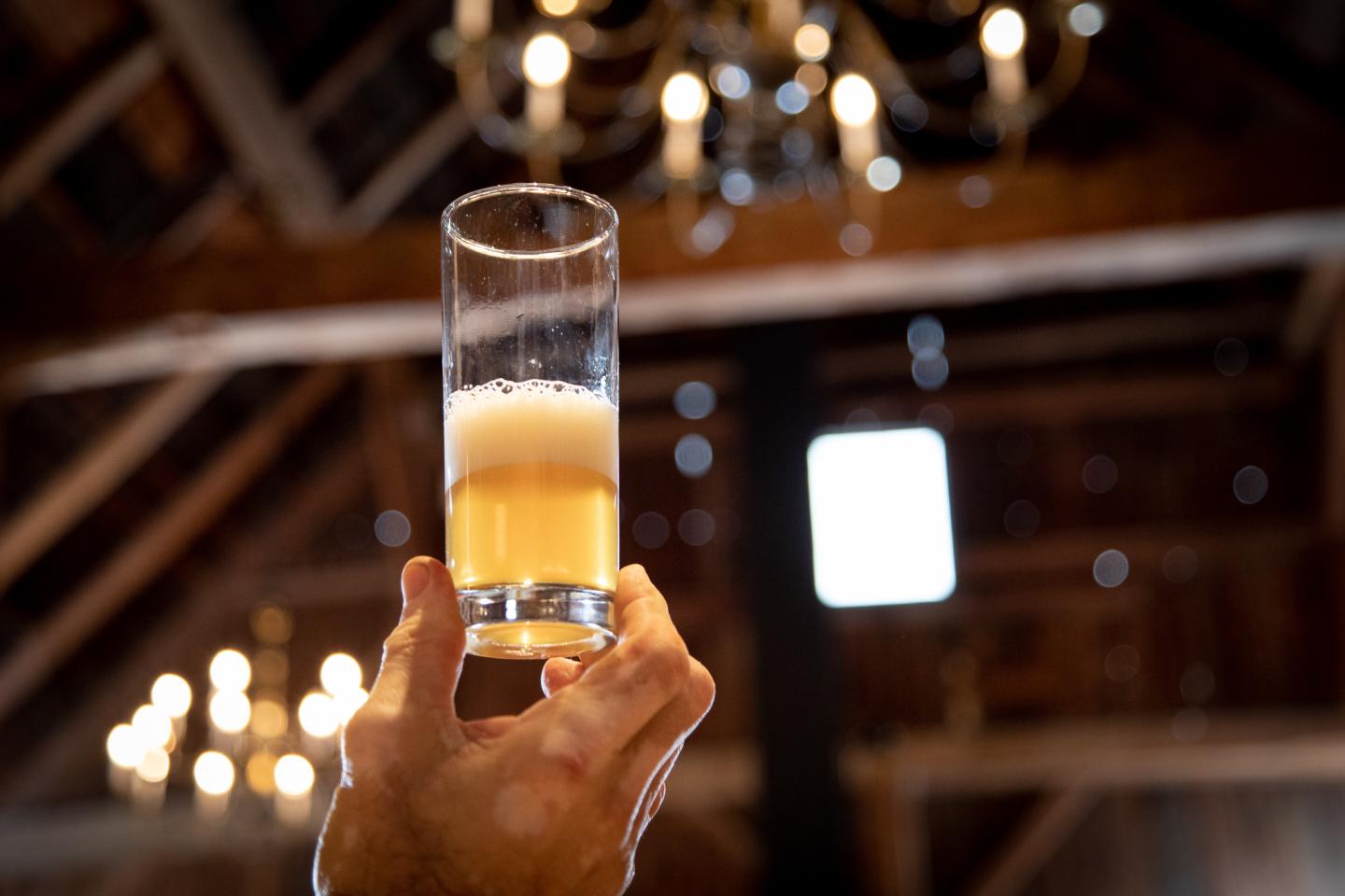 Hand holding a tall glass of beer in a dim, chandelier-lit room.
