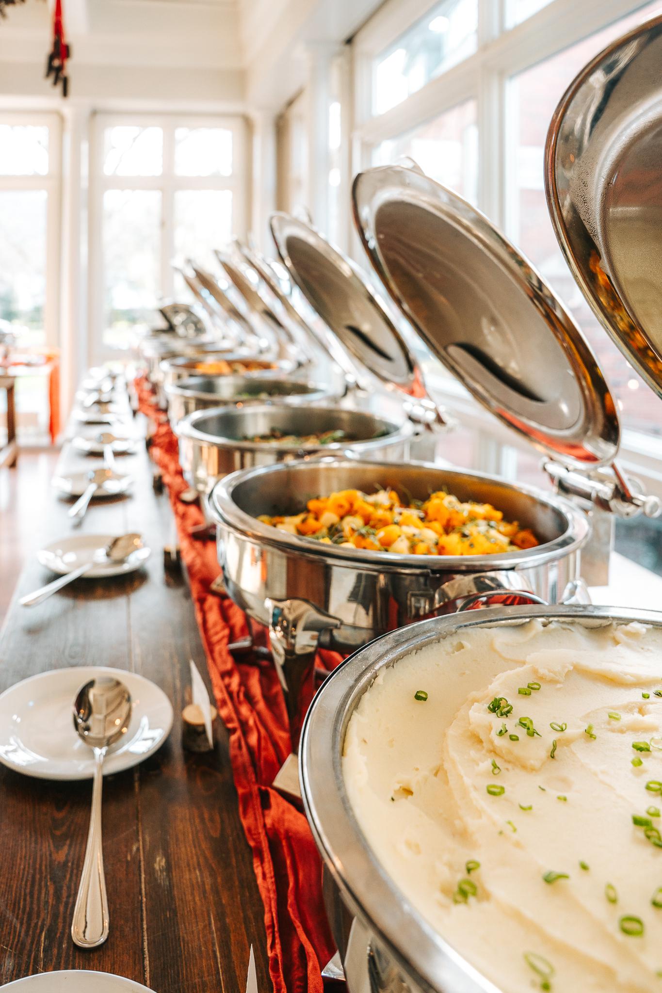 Buffet table with chafing dishes of varied foods near large windows.