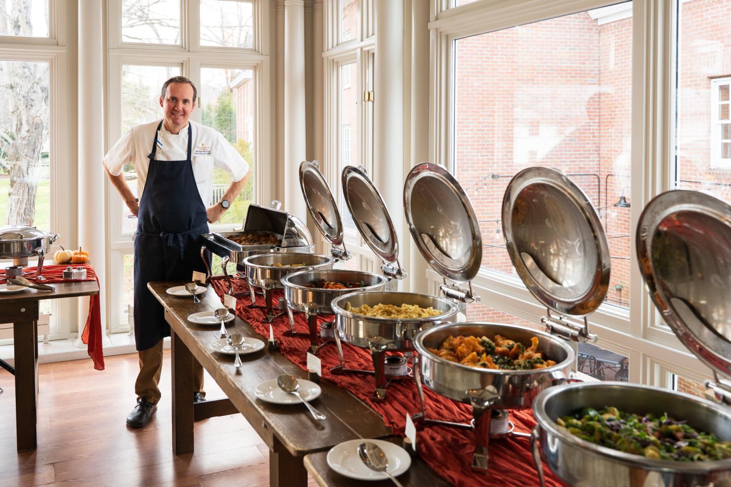 Chef standing by a buffet table with various dishes in a bright room.