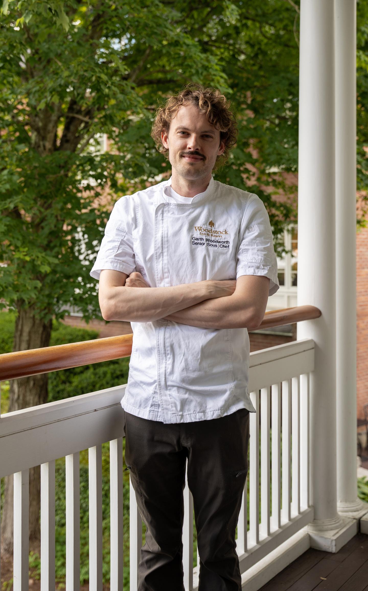Woodstock Inn Chef Garth on a porch, arms crossed, with greenery behind.