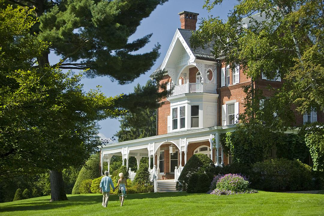 Victorian house with large porch, surrounded by trees and green lawn.